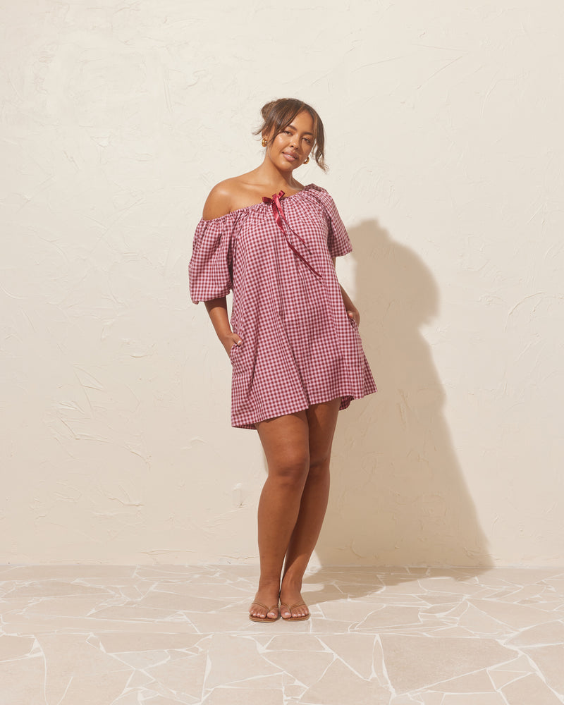 FLORY MINI DRESS PINK BURGUNDY GINGHAM | Cotton mini dress designed with a gathered neckline with a ribbon at the centre front. Made from organic cotton in a burgundy and pink gingham, this dress can be worn...