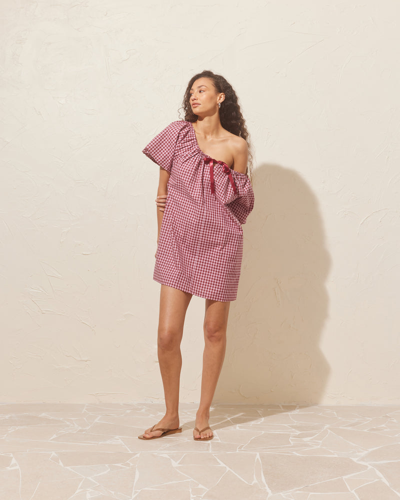 FLORY MINI DRESS PINK BURGUNDY GINGHAM | Cotton mini dress designed with a gathered neckline with a ribbon at the centre front. Made from organic cotton in a burgundy and pink gingham, this dress can be worn...